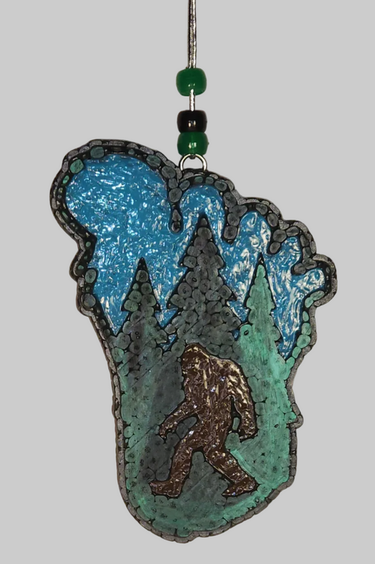 A footprint shaped car air freshener featuring a black silhouette of bigfoot against a blue background with green trees.