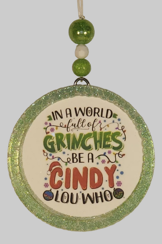 A round car air freshener with a sparkly green and multicolored design, featuring the text: 'In a world full of Grinches, be a Cindy Lou Who"